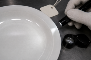Close-up ceramic dinnerware crazing inspection with fine glaze cracks, flashlight, magnifier, and sample tag