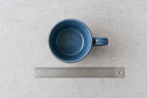 Overhead view of a blue ceramic mug beside a ruler, showing why the handle belongs in the overall width record