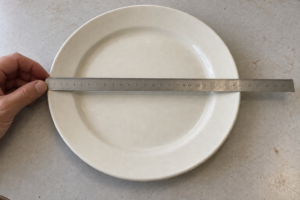 A hand steadies a steel ruler across the center of an ivory ceramic plate to illustrate an outside diameter check