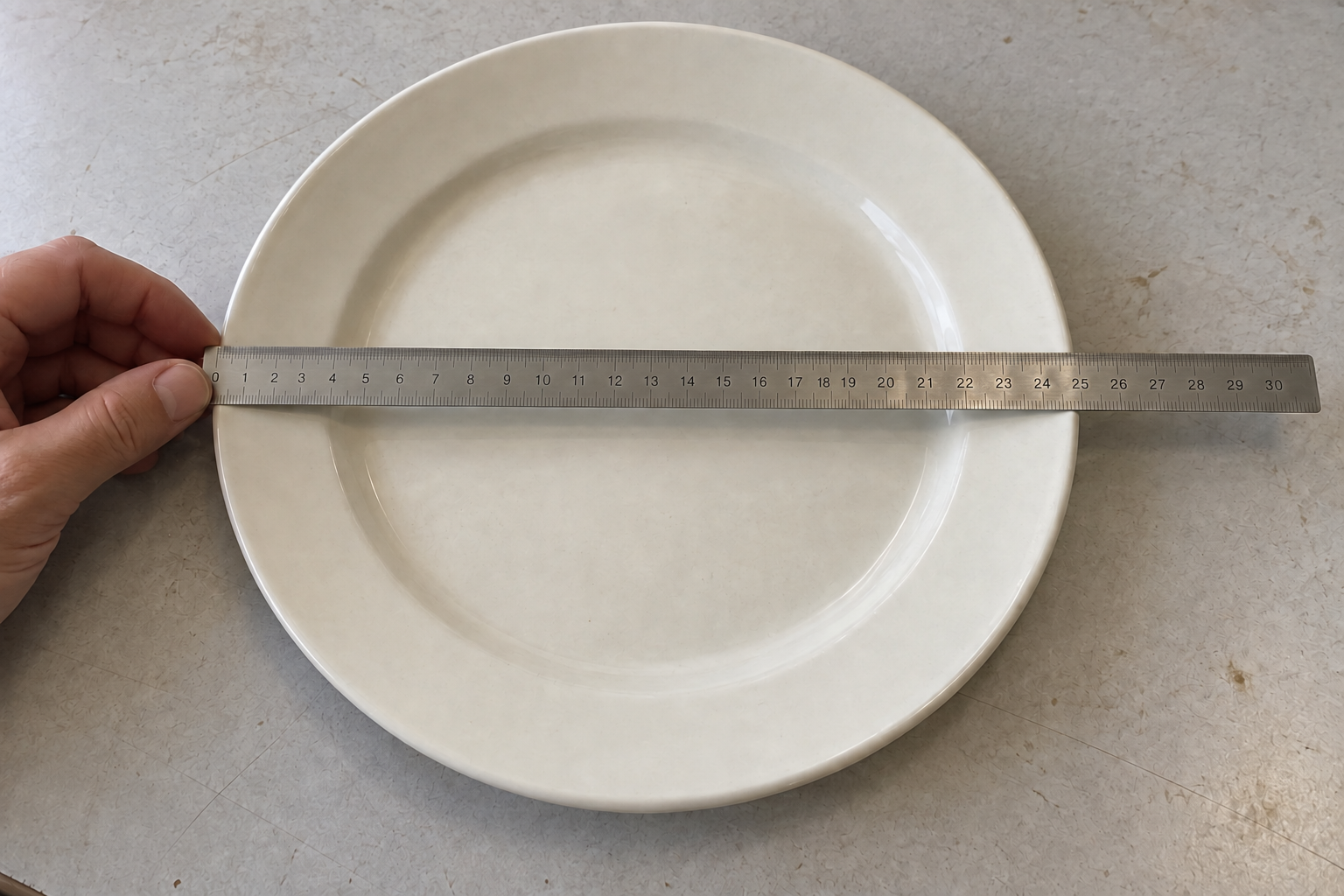 A hand steadies a steel ruler across the center of an ivory ceramic plate to illustrate an outside diameter check