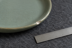 Close view of a small chip on a sage ceramic plate rim resting on a padded inspection surface beside a plain ruler