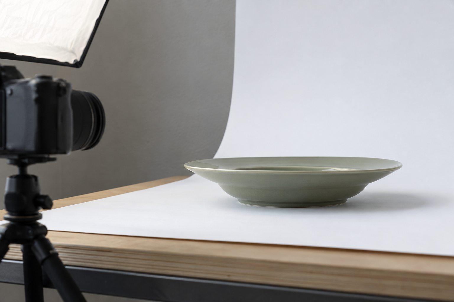 Side view of a wide-rim sage ceramic bowl on a white photo sweep, with a camera set low to show its shallow profile