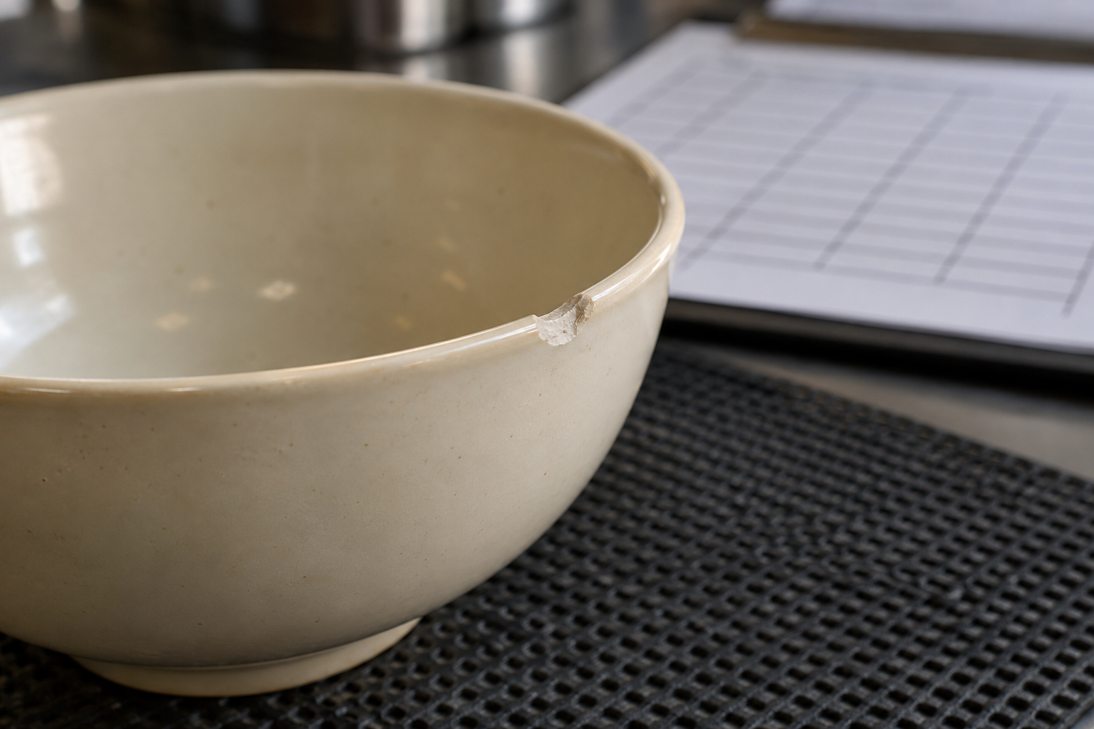 Close view of a small chip on a cream ceramic bowl rim under inspection light, with a plain stock tally sheet behind