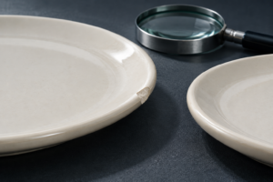 Close view of a chipped ivory ceramic plate rim beside an intact matching edge on a laboratory inspection mat.