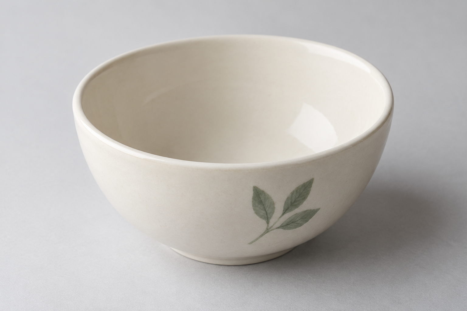 Angled close view of a ceramic bowl with a smooth cream interior and a simple green leaf motif on its outer wall