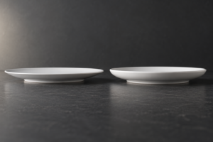 Low side view of two white porcelain plates showing different rim profiles and foot rings on a dark comparison surface
